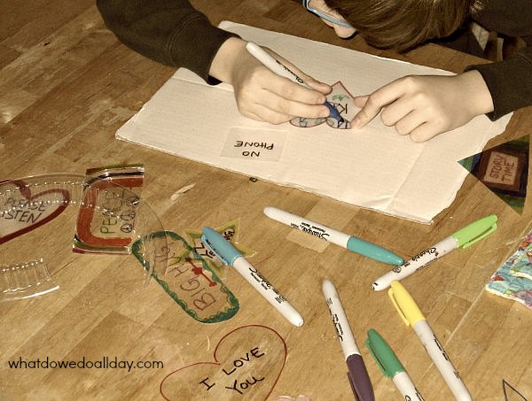Child Making kindness tokens for a family harmony project by drawing on a plastic heart surrounded by art project materials.
